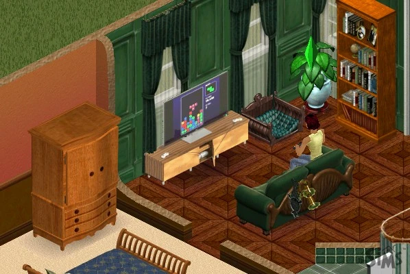 A flat panel on a wooden media console playing a falling-block puzzle game, with a Sim on a green sofa holding a controller.